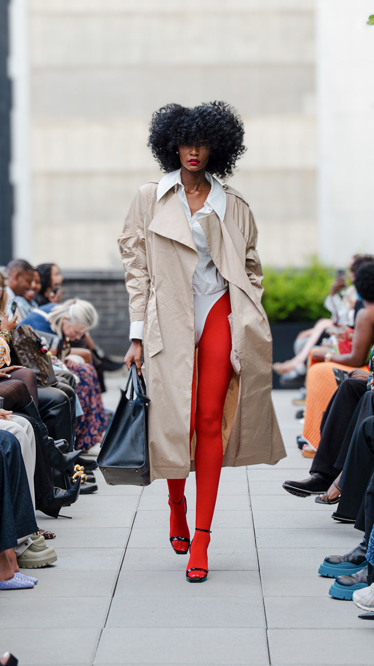 Kylar Smith walking on a catwalk wearing a beige trench coat, white bodysuit, red tights, and black heels.