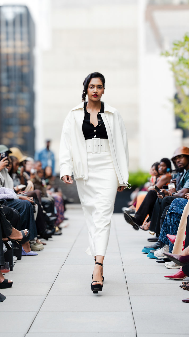 Arifah Akther walking on Megan Renee's SS26 fashion show runway wearing a white vegan leather jacket and skirt set.